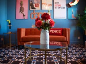 A room with a red sofa and blue walls with interesting pictures of all colours on them, and a patterned carpet. There is a vase of red and pink flowers in the foreground.