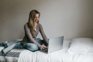 Woman sitting cross legged on a white bed. She is typing on a computer that is on the bed in front of her