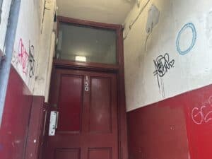 Passageway painted cream and red with lots of graffiti on it, with a badly maintained door with badly spaced numbers.